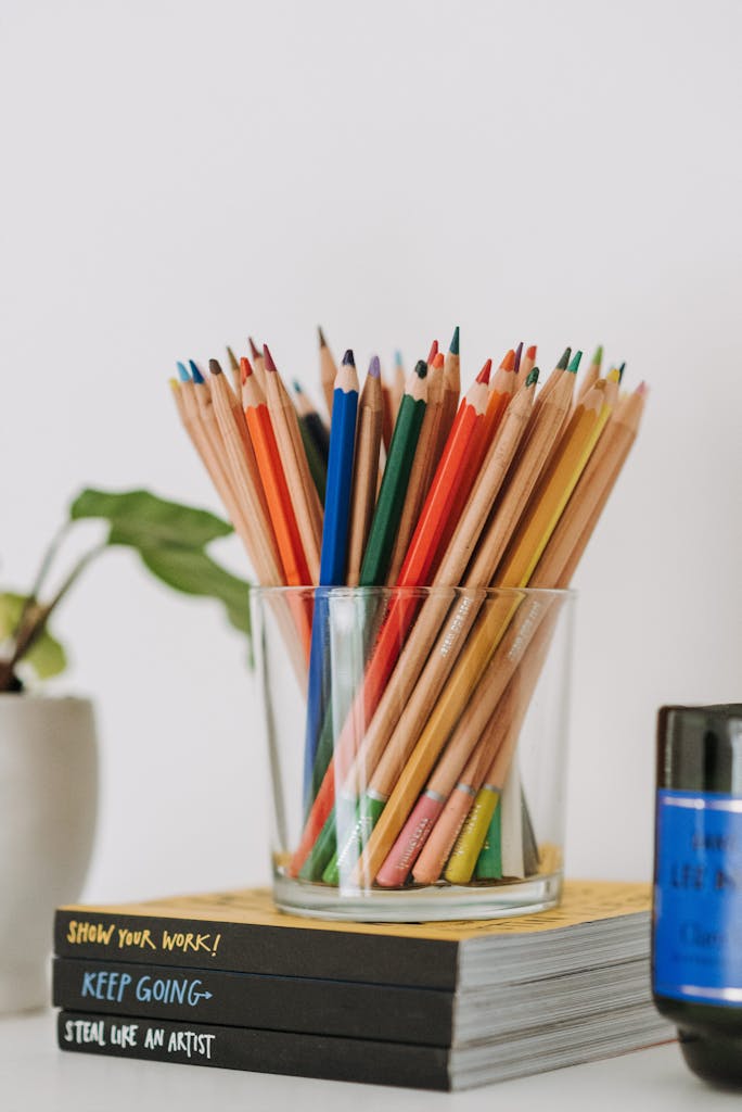 Set of various multicolored pencils in glass cup placed on stack of books in light room with green plant on white background